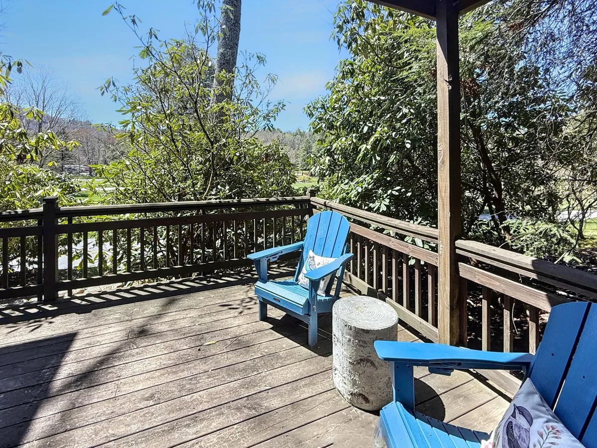 Two blue Adirondack chairs and a small table rest on an exterior deck enveloped by greenery. The scene hints at quiet moments spent reading, socializing, or just enjoying the beauty and serenity of the outdoors.