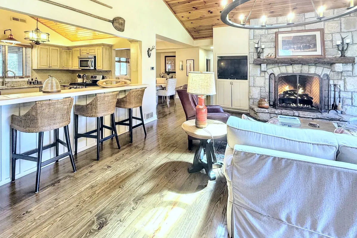 This image showcases a spacious open-concept great room with a prominent custom stone fireplace, comfortable seating, visible kitchen island, wooden ceiling beams, and a cozy breakfast nook. The scene exudes warmth and encourages gatherings, with multiple elements of the living, dining, and kitchen spaces visible in one frame.