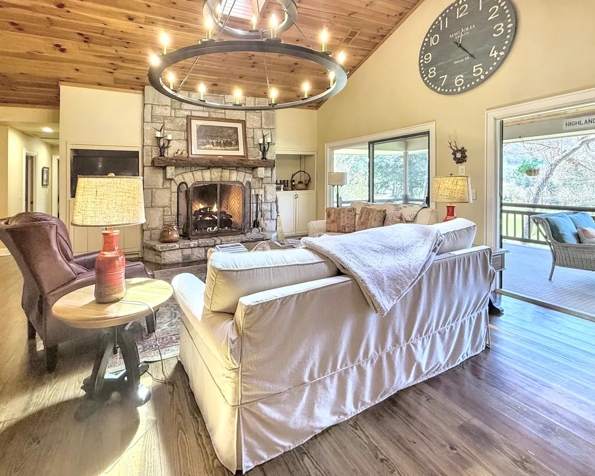 A spacious great room centered around a striking stone fireplace, surrounded by comfortable seating, a chandelier overhead, and a large clock as a focal point. The inviting atmosphere encourages gathering and relaxation after a productive day.