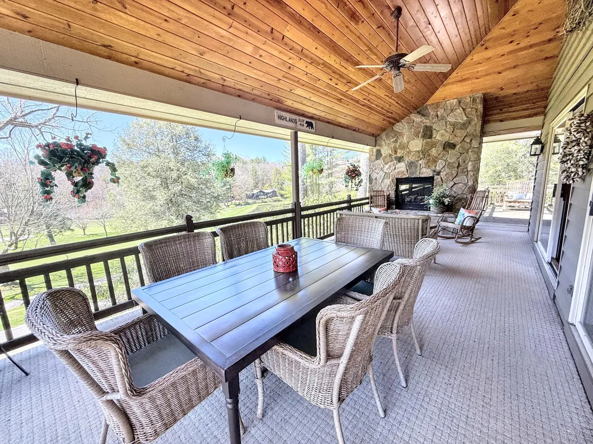 A spacious covered patio featuring a large dining table, hanging plants, a prominent stone gas fireplace, and a wooden ceiling. The setup is arranged for both dining and relaxing, with ample space for gatherings and cozy evenings by the fire. The background suggests an inviting view out to the mountains or golf course.