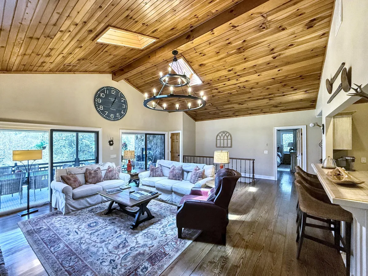 An airy living area with a large white sofa, exposed wooden beams, a chandelier, and plenty of natural light, creating an inviting space for relaxation and socializing after work. Decorative touches and a visible deck hint at scenic surroundings.