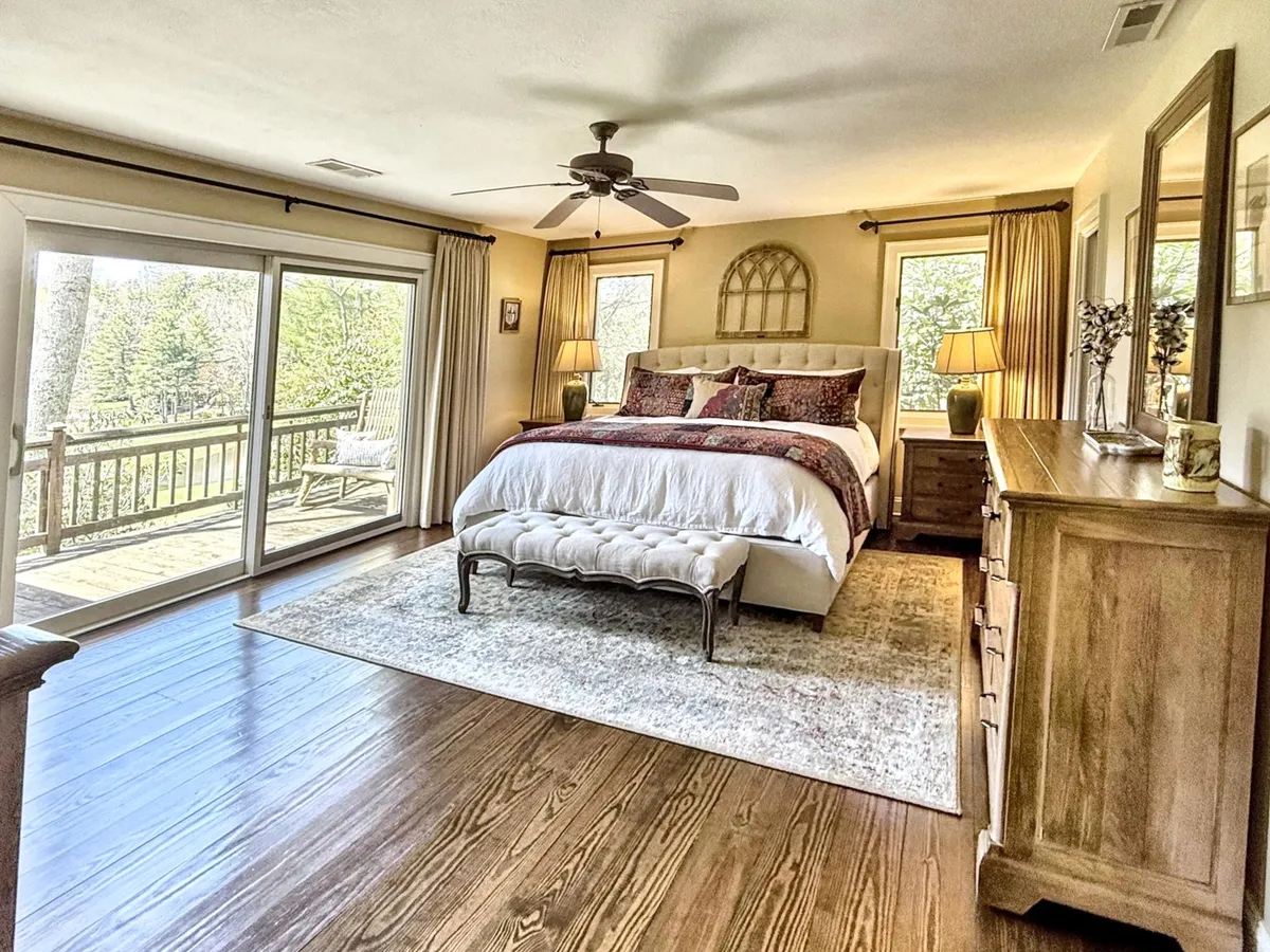 A serene master bedroom showcasing a spacious bed adorned with decorative pillows, large windows offering abundant natural light and beautiful views, a wooden dresser, and a cozy area rug. The ambiance is peaceful, inviting restful relaxation and private retreat.