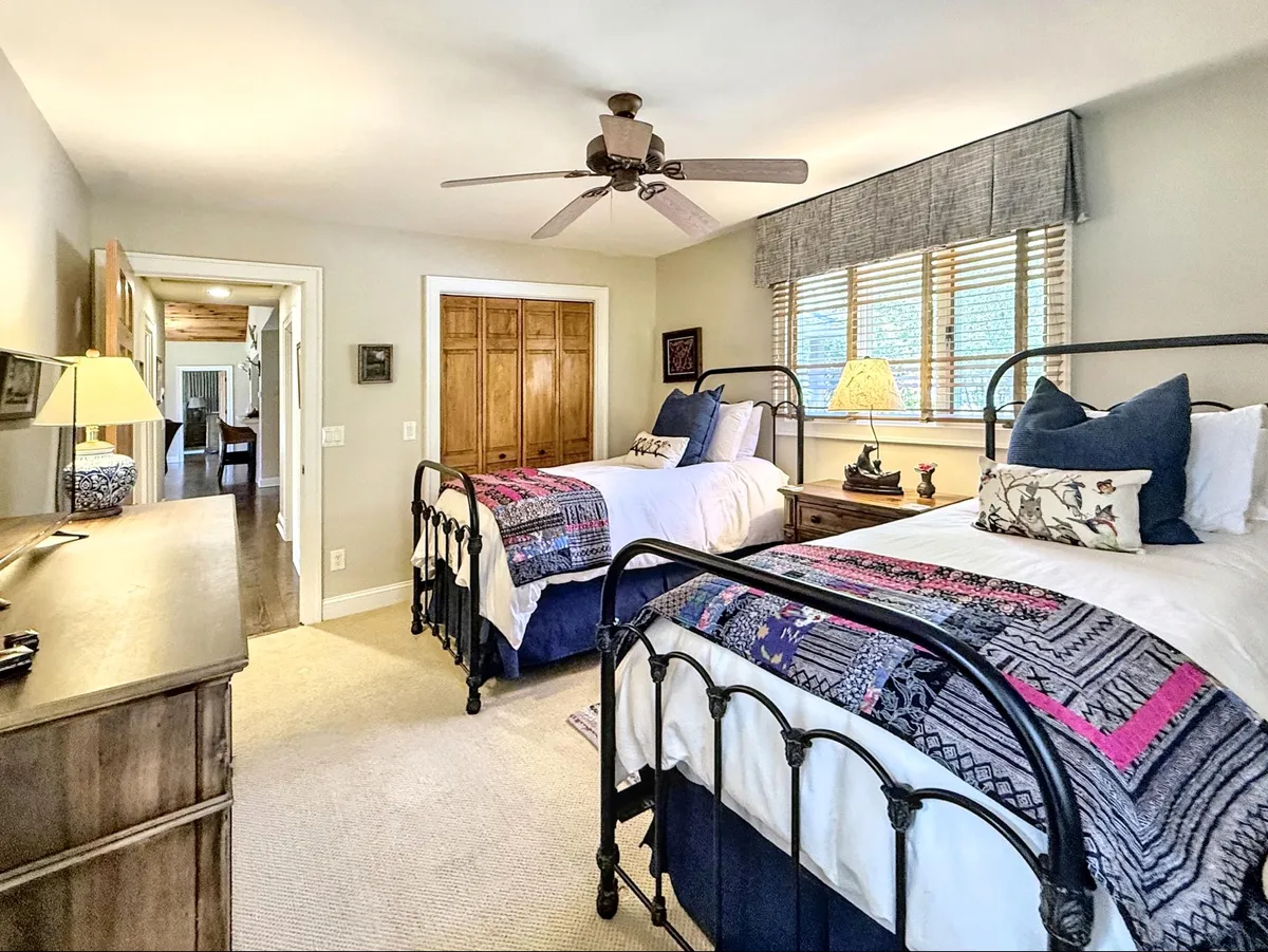 A cozy twin bedroom featuring two metal-framed beds with vibrant blankets, a wooden dresser with a lamp, and generous natural light streaming through large windows. The room feels both inviting and functional, ideal for families or friends seeking separation and comfort.